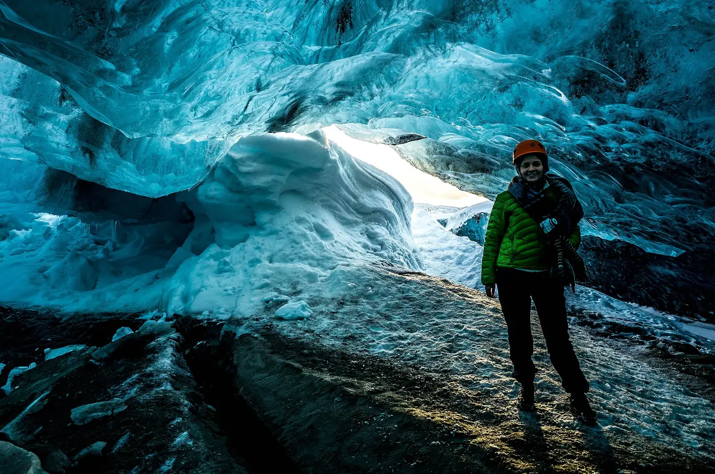A visitor in a helmet at the bright exit of a blue glacier ice cave in Iceland at the end of an Iceland ice cave day tour